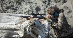 A young woman using a dead tree to brace her hunting rifle